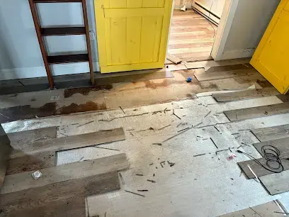 Lifted floorboards on a wet floor near a yellow door and a stepladder in a Wareham cleanup
