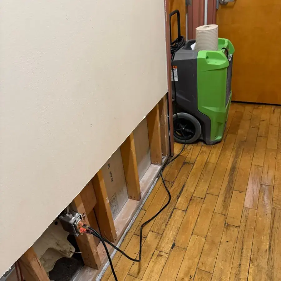 Drywall cut away above a hardwood floor with a dehumidifier running beside the wall
